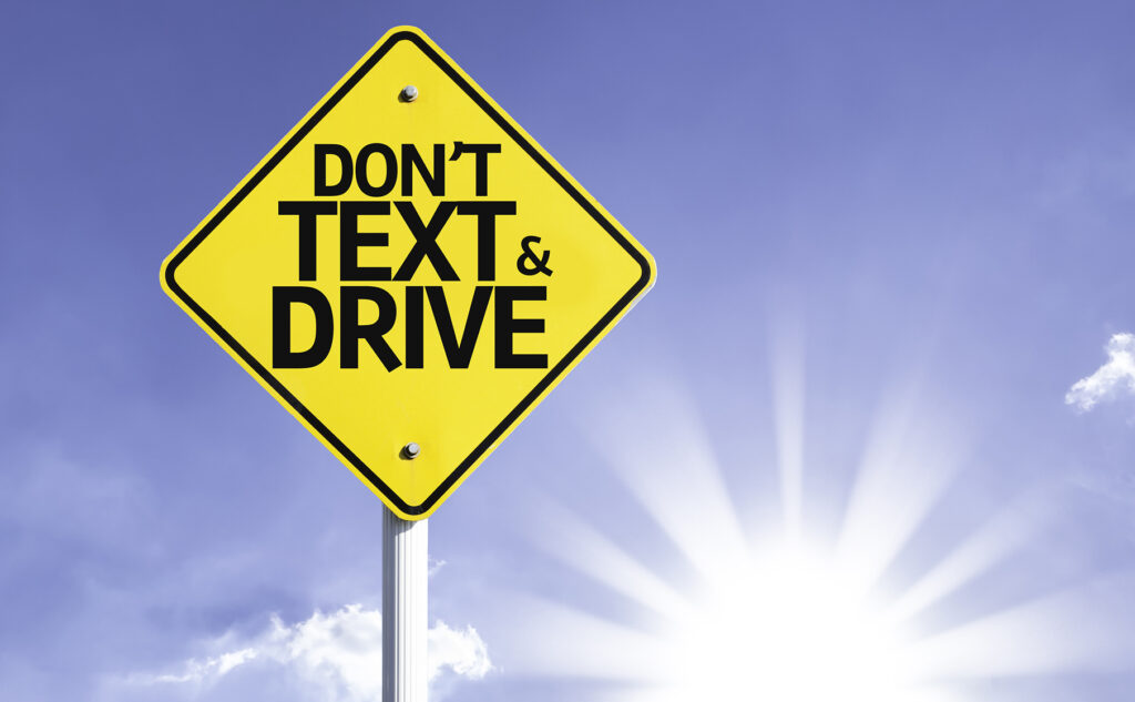 A yellow image of a "Don't Text & Drive" sign against a backdrop of the sky and the sun.