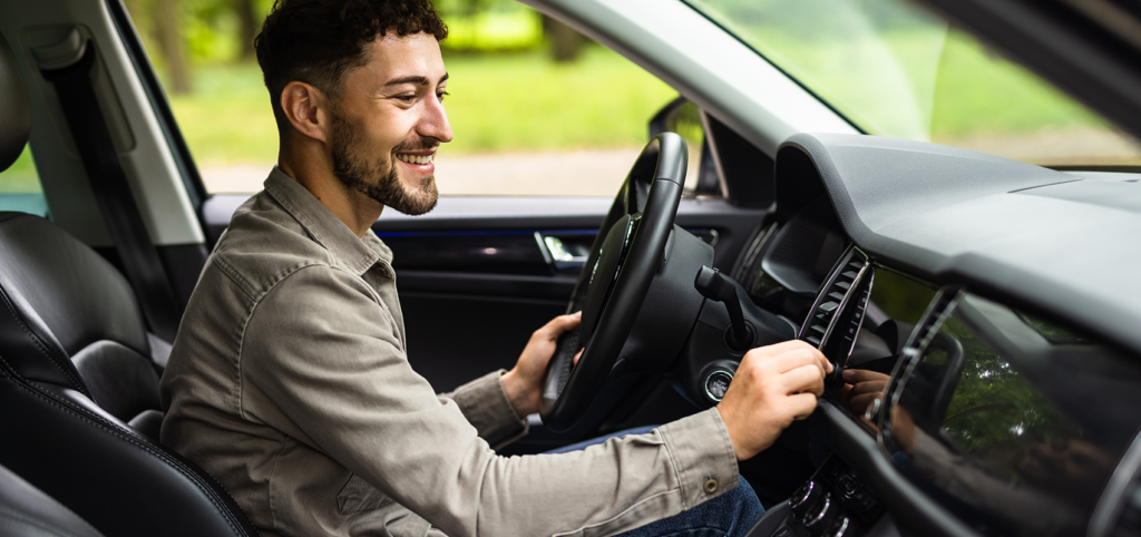 Smiling young man adjusting the infotainment screen inside a modern car while holding the steering wheel — the ultimate playlist for every drive
